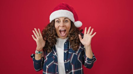 Foto de Mujer hispana de mediana edad expresión sorpresa usando sombrero de Navidad sobre fondo rojo aislado - Imagen libre de derechos