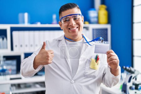 Téléchargez les photos : Jeune homme hispanique travaillant au laboratoire scientifique tenant carte d'identité souriant heureux et positif, pouce levé faire excellent et signe d'approbation - en image libre de droit
