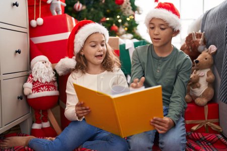 Foto de Dos niños leyendo libro sentado en el suelo por el árbol de Navidad en casa - Imagen libre de derechos