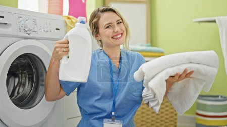 Téléchargez les photos : Jeune femme blonde nettoyant professionnel tenant bouteille de détergent et serviettes pliées à la buanderie - en image libre de droit