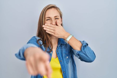 Téléchargez les photos : Jeune femme blonde debout sur fond bleu se moquant de vous, pointant du doigt la caméra avec la main sur la bouche, expression de honte - en image libre de droit