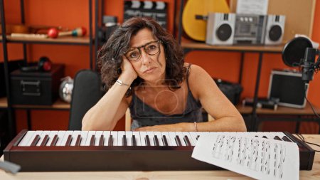Foto de Músico hispano de mediana edad apoyado en el piano con expresión triste en el estudio de música - Imagen libre de derechos