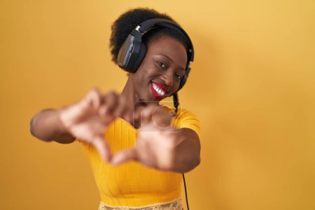 Téléchargez les photos : Femme africaine avec les cheveux bouclés debout sur fond jaune portant des écouteurs souriant dans l'amour faisant forme de symbole de coeur avec les mains. concept romantique. - en image libre de droit