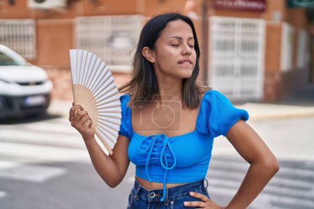 Foto de Mujer hispana joven usando ventilador de mano sudando en la calle - Imagen libre de derechos