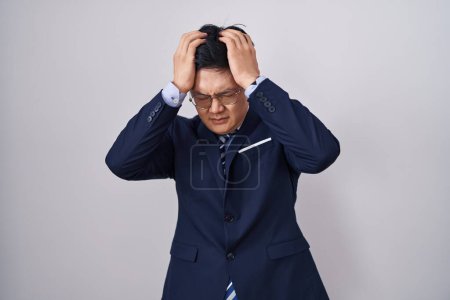 Foto de Joven hombre asiático vistiendo traje de negocios y corbata sufriendo de dolor de cabeza desesperado y estresado porque el dolor y la migraña. manos en la cabeza. - Imagen libre de derechos