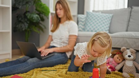 Téléchargez les photos : Mère caucasienne sérieuse télétravail sur ordinateur portable, ainsi que la petite fille détendue jouer avec poupée sur couverture de sol dans le salon à la maison - en image libre de droit