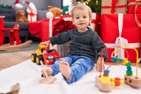 Photo for Adorable blond toddler playing with toys sitting on floor by christmas gifts at home - Royalty Free Image