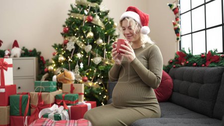 Foto de Mujer embarazada joven oliendo taza de café celebrando la Navidad en casa - Imagen libre de derechos