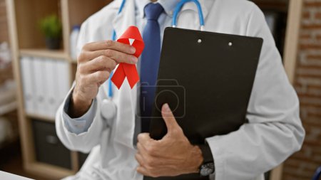 Téléchargez les photos : Jeune homme hispanique médecin tenant ruban rouge et presse-papiers à la clinique - en image libre de droit