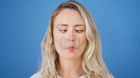 Téléchargez les photos : Jeune femme blonde souriante confiante faire geste drôle avec la bouche sur fond bleu isolé - en image libre de droit