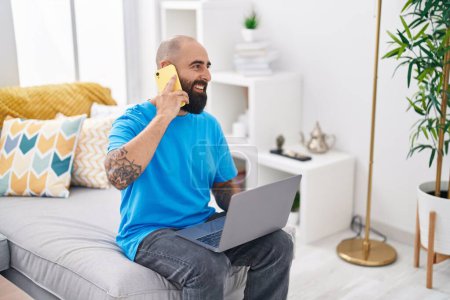 Photo for Young bald man talking on smartphone using laptop at home - Royalty Free Image