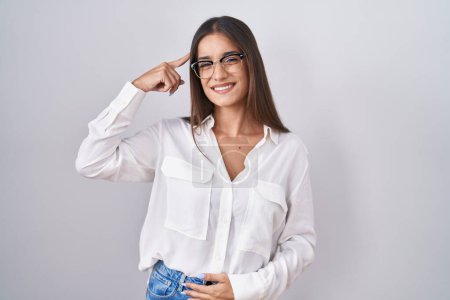 Téléchargez les photos : Jeune femme brune portant des lunettes souriant pointant vers la tête avec un doigt, bonne idée ou pensée, bonne mémoire - en image libre de droit