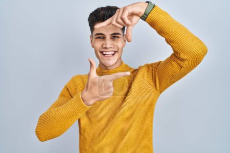Foto de Joven hombre hispano de pie sobre fondo azul sonriendo haciendo marco con manos y dedos con cara feliz. concepto de creatividad y fotografía. - Imagen libre de derechos