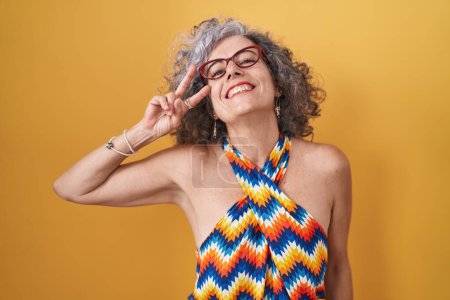 Photo for Middle age woman with grey hair standing over yellow background doing peace symbol with fingers over face, smiling cheerful showing victory - Royalty Free Image