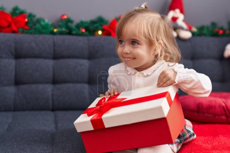 Photo for Adorable blonde girl unpacking gift sitting on sofa by christmas decor at home - Royalty Free Image