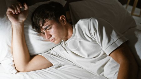 Téléchargez les photos : Épuisé jeune homme hispanique trouve le confort dans un sommeil matinal lumineux, couché sans être dérangé dans sa chambre confortable, cédant à l'étreinte douce de son lit et oreiller, dans un portrait intérieur détendu. - en image libre de droit