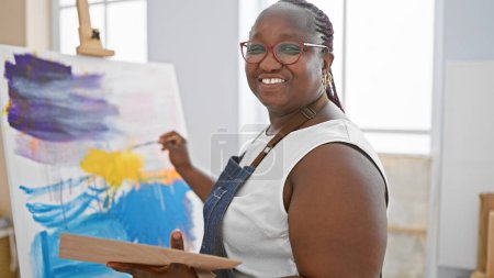 Foto de Mujer africana americana confiada y sonriente dibujando alegremente en un estudio de arte con su pincel y lienzo, abrazando la creatividad. - Imagen libre de derechos