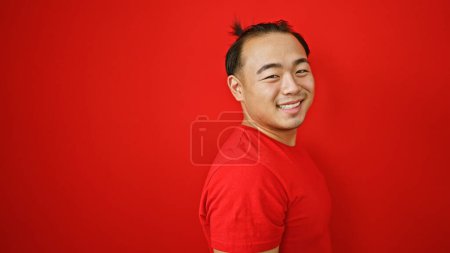 Foto de Joven hombre chino alegre, confiado y riendo, parado sobre fondo rojo aislado, exudando felicidad y energía positiva - Imagen libre de derechos