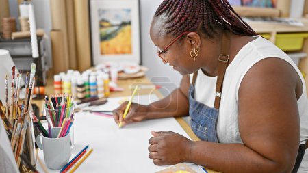 Téléchargez les photos : Femme afro-américaine focalisée, artiste captivante, dessinant avec concentration sur son carnet dans un studio d'art d'intérieur. - en image libre de droit