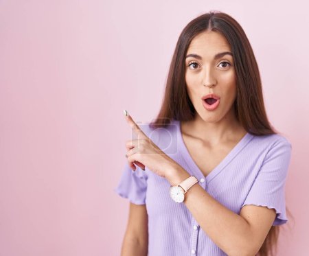 Photo for Young hispanic woman with long hair standing over pink background surprised pointing with finger to the side, open mouth amazed expression. - Royalty Free Image