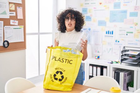Photo for Young middle east woman holding recycling bag with plastic bottles at the office clueless and confused expression. doubt concept. - Royalty Free Image