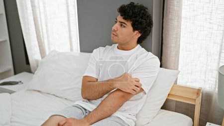 Téléchargez les photos : Jeune homme latino bras à gratter pour démangeaisons assis sur le lit dans la chambre - en image libre de droit