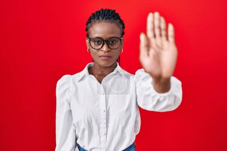Foto de Mujer africana con trenzas de pie sobre fondo rojo haciendo dejar de cantar con la palma de la mano. expresión de advertencia con gesto negativo y grave en la cara. - Imagen libre de derechos