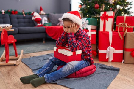 Téléchargez les photos : Adorable garçon hispanique tenant cadeau de Noël assis sur le sol avec une expression malheureuse à la maison - en image libre de droit