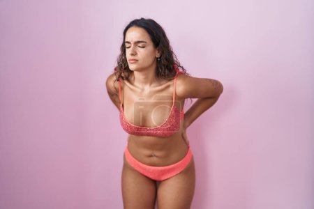 Foto de Mujer hispana joven vistiendo lencería sobre fondo rosa sufriendo de dolor de espalda, tocando con la mano, dolor muscular - Imagen libre de derechos
