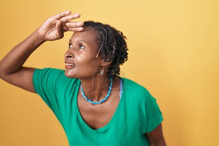 Photo for African woman with dreadlocks standing over yellow background very happy and smiling looking far away with hand over head. searching concept. - Royalty Free Image