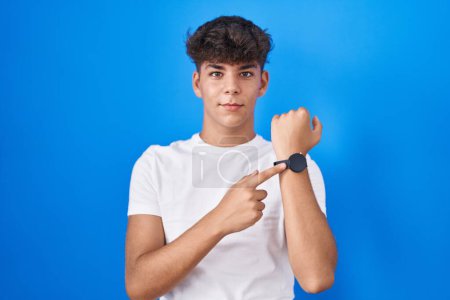 Téléchargez les photos : Adolescent hispanique debout sur fond bleu pressé pointant vers regarder le temps, l'impatience, regardant la caméra avec une expression détendue - en image libre de droit