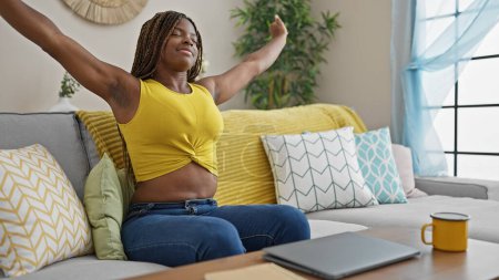 Téléchargez les photos : Femme afro-américaine assise sur un canapé étirant les bras fatigué à la maison - en image libre de droit