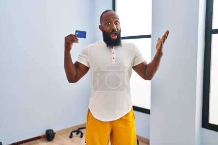 Téléchargez les photos : Homme afro-américain portant des vêtements de sport tenant la carte de crédit effrayé et étonné avec la bouche ouverte pour la surprise, visage incrédule - en image libre de droit