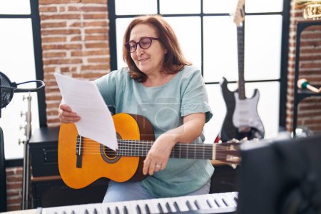 Photo for Senior woman musician playing classical guitar reading paper at music studio - Royalty Free Image