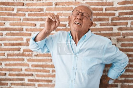Téléchargez les photos : Homme âgé avec les cheveux gris debout sur le mur de briques étirant le dos, fatigué et détendu, somnolent et bâillant pour tôt le matin - en image libre de droit