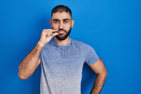 Téléchargez les photos : Moyen-Orient homme avec barbe debout sur fond bleu bouche et lèvres fermées comme zip avec les doigts. secret et silencieux, tabou parlant - en image libre de droit