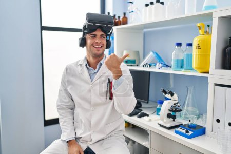 Téléchargez les photos : Jeune homme hispanique travaillant au laboratoire scientifique portant des lunettes vr pointant le pouce vers le côté souriant heureux avec la bouche ouverte - en image libre de droit