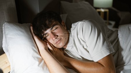 Foto de Un joven hispano agotado encuentra consuelo en un brillante sueño matutino, acostado tranquilo en su acogedor dormitorio, entregándose al suave abrazo de su cama y almohada, en un retrato interior relajado. - Imagen libre de derechos