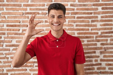 Téléchargez les photos : Jeune homme hispanique debout au-dessus du mur de briques geste souriant et confiant avec la main faisant signe de petite taille avec les doigts regardant et la caméra. concept de mesure. - en image libre de droit