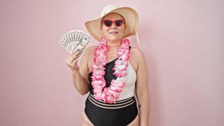 Foto de Turista mujer de pelo gris de mediana edad con traje de baño y lei hawaiano sosteniendo dólares sobre un fondo rosa aislado - Imagen libre de derechos