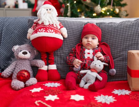 Photo for Adorable caucasian baby holding teddy bear sitting on sofa by christmas tree at home - Royalty Free Image