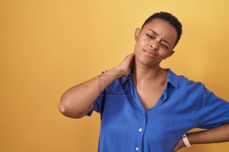 Téléchargez les photos : Femme afro-américaine debout sur fond jaune souffrant d'une blessure au cou, toucher le cou avec la main, douleur musculaire - en image libre de droit