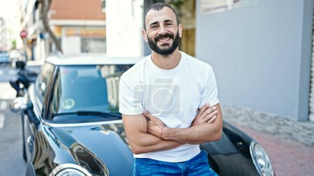 Téléchargez les photos : Jeune homme hispanique souriant confiant assis sur la voiture avec les bras croisés geste à la rue - en image libre de droit