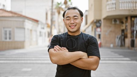 Téléchargez les photos : Jeune homme chinois joyeux, rayonnant de confiance et de joie, repéré souriant et debout les bras croisés dans une rue urbaine animée - en image libre de droit