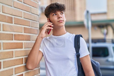 Téléchargez les photos : Jeune adolescent hispanique parlant sur smartphone avec une expression détendue dans la rue - en image libre de droit