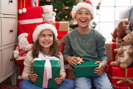 Téléchargez les photos : Deux enfants tenant un cadeau assis sur le sol près de l'arbre de Noël à la maison - en image libre de droit