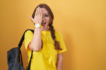 Téléchargez les photos : Jeune femme caucasienne portant un sac à dos étudiant sur fond jaune couvrant un oeil avec la main, sourire confiant sur le visage et l'émotion surprise. - en image libre de droit