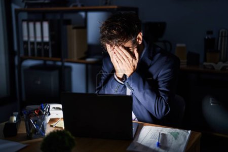 Foto de Joven hispano que trabaja en la oficina por la noche con expresión triste cubriendo la cara con las manos mientras llora. concepto de depresión. - Imagen libre de derechos