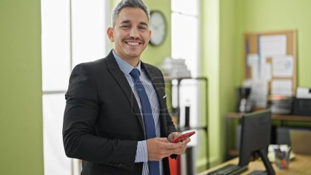 Foto de Trabajador de negocios joven hispano sonriendo confiado usando teléfono inteligente en la oficina - Imagen libre de derechos