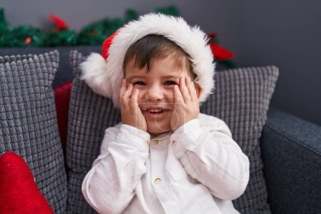 Photo for Adorable hispanic toddler wearing christmas hat sitting on sofa at home - Royalty Free Image